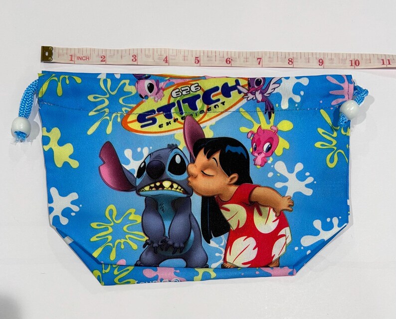 May include: Blue drawstring pouch featuring a graphic of Stitch and Lilo from the animated film. The pouch has a blue background with yellow and white splatters. The text "626 Experiment" is printed on the pouch. The pouch is approximately 10 inches wide.