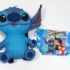 May include: A plush blue Stitch character with large eyes and ears, next to a blue drawstring bag featuring Stitch and Lilo kissing. The bag has a floral pattern and the text "Stitch".