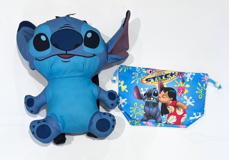 May include: A plush blue Stitch character with large eyes and ears, next to a blue drawstring bag featuring Stitch and Lilo kissing. The bag has a floral pattern and the text "Stitch".