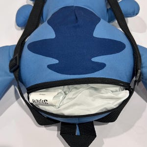 May include: A blue Stitch-shaped backpack with a black zipper and shoulder strap. The backpack features a dark blue design on the front and the word "Stitch" printed on a white tag inside the open compartment.
