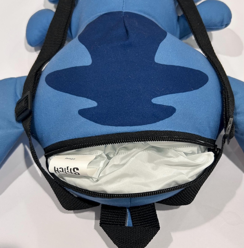 May include: A blue Stitch-shaped backpack with a black zipper and shoulder strap. The backpack features a dark blue design on the front and the word "Stitch" printed on a white tag inside the open compartment.
