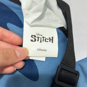 May include: A close-up of a blue bag with a black strap and buckle. The bag has a white tag with the Disney Stitch logo and the Disney copyright symbol.