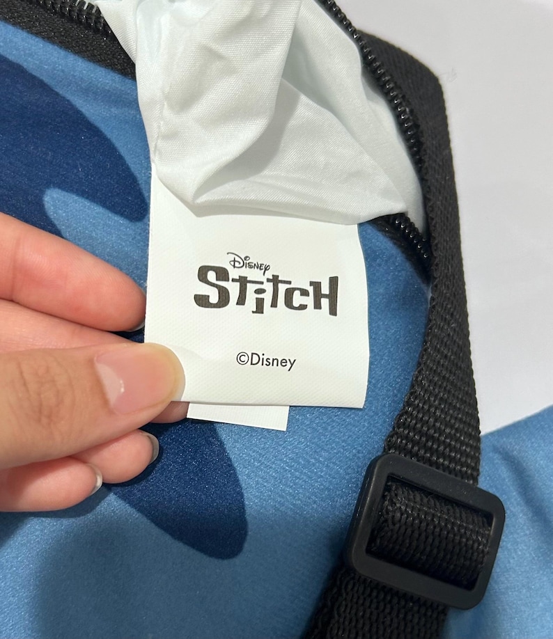 May include: A close-up of a blue bag with a black strap and buckle. The bag has a white tag with the Disney Stitch logo and the Disney copyright symbol.