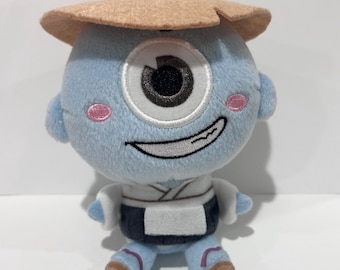 May include: A blue plush toy of a cyclops-like character wearing a tan hat. The toy has a large eye, pink cheeks, and a smiling mouth. It is dressed in a white and black outfit, with small arms and legs.