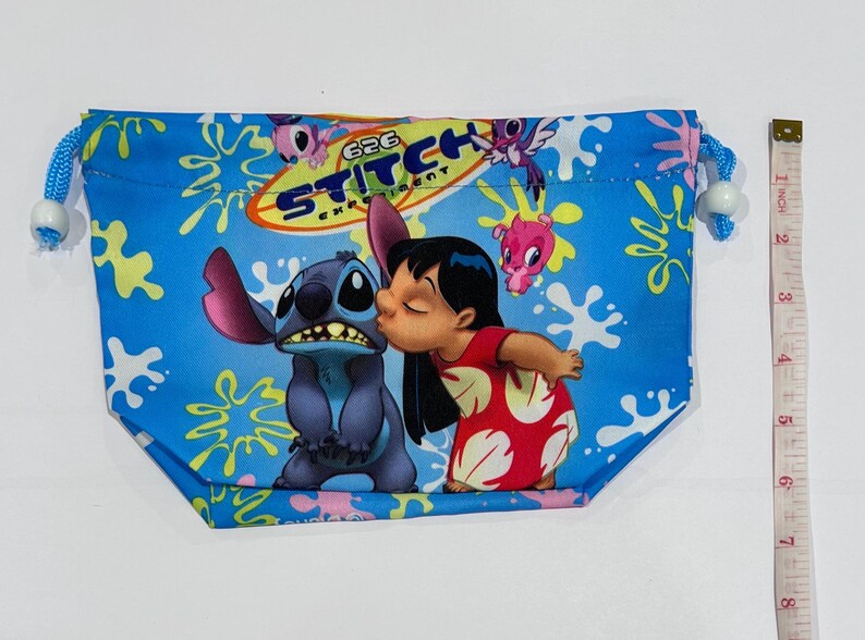 May include: A blue drawstring bag featuring Stitch and Lilo from the animated film Lilo & Stitch. The bag has a blue background with white splatters and the words "626 Experiment Stitch". The bag is approximately 8 inches wide.