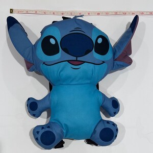 May include: A blue plush backpack shaped like the character Stitch from Lilo & Stitch. The backpack has large ears, big eyes, and a smiling face. The backpack measures approximately 15 inches in length.
