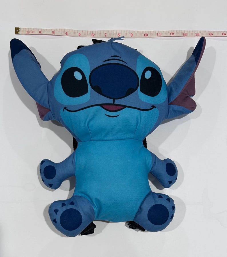 May include: A blue plush backpack shaped like the character Stitch from Lilo & Stitch. The backpack has large ears, big eyes, and a smiling face. The backpack measures approximately 15 inches in length.