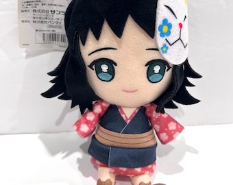 Op de afbeelding: Een pluche speelgoed van een personage met zwart haar, dat een roze en witte kimono draagt met een wit masker met blauwe bloemen. Het personage heeft een glimlachend gezicht met blauwe ogen.