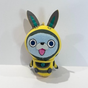 May include: A plush toy featuring a cartoon character with a yellow and black bee-themed outfit. The character has a blue face, large blue eyes, and a pink tongue. It has long, gray and green bunny ears and a small chain for hanging.