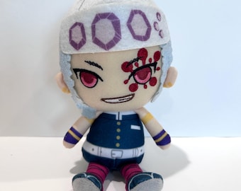 Op de afbeelding: Een pluche pop van een personage met een lichte huid, rode ogen en een witte hoofdbedekking met paarse accenten. De pop draagt een donkerblauwe outfit met een witte riem, roze en blauw gestreepte sokken en grijze schoenen. Het personage heeft een rode vlek op het gezicht.