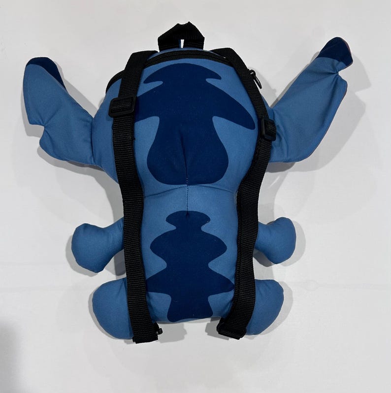 May include: A blue Stitch-shaped backpack with black adjustable straps. The backpack features a dark blue center and lighter blue arms, legs, and ears. The backpack has a top handle and a zipper closure.