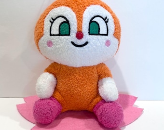May include: A plush toy character with an orange body, white face, and green eyes. The toy has pink cheeks and feet, and sits on a pink felt flower. A red loop is attached to the top.