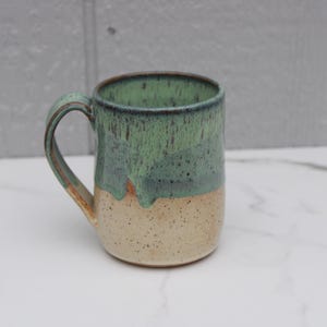 May include: A handmade ceramic mug with a rounded body and a large handle. The mug features a two-tone design, with a speckled beige base and a green and blue glazed top. The mug is approximately 4 inches tall.