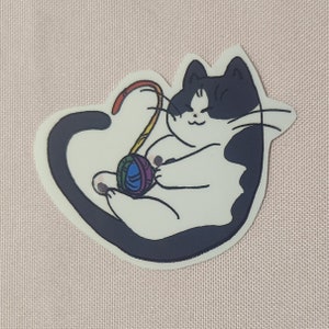 Könnte beinhalten: Eine schwarz-weiße Katze mit einem Regenbogen-Wollknäuel. Die Katze sitzt auf ihrem Schwanz und hat einen glücklichen Gesichtsausdruck.