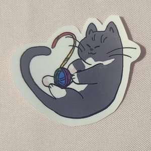 Könnte beinhalten: Ein Aufkleber mit einer grauen Katze mit weißen Abzeichen, die mit einem regenbogenfarbenen Wollknäuel spielt. Die Katze ist zusammengerollt, mit einem zufriedenen Ausdruck, und die Wolle bildet eine gebogene Form.