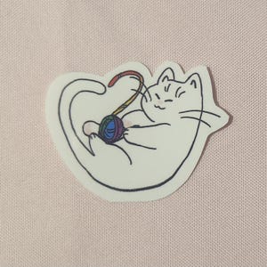 Könnte beinhalten: Ein weißer Katzenaufkleber mit einem Regenbogen-Garnball. Die Katze liegt auf der Seite mit dem Schwanz um den Körper gewickelt. Die Katze hat einen glücklichen Gesichtsausdruck.