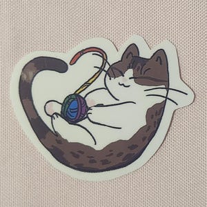 Könnte beinhalten: Eine braune und weiße Katze mit einem Regenbogen-Wollknäuel. Die Katze ist zu einem Kreis zusammengerollt, mit dem Schwanz um den Körper gewickelt. Die Katze schaut mit einem glücklichen Ausdruck auf das Wollknäuel.