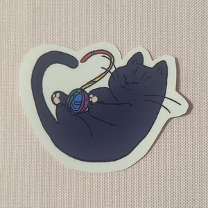Könnte beinhalten: Eine schwarze Katze spielt mit einem bunten Wollknäuel. Die Katze liegt auf der Seite, der Schwanz ist um den Körper gewickelt. Die Wolle ist regenbogenfarben und die Katze schaut auf die Wolle.