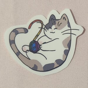 Könnte beinhalten: Ein Cartoon-Aufkleber eines weißen Katers mit grauen und braunen Flecken, der mit einem bunten Wollknäuel spielt. Die Katze hat einen glücklichen Ausdruck und ist in einer verspielten Pose zusammengerollt.