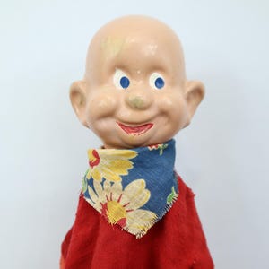 May include: Vintage doll with a bald head, blue eyes, and a smiling expression. The doll wears a red outfit and a blue scarf with yellow floral patterns. A classic collectible toy.