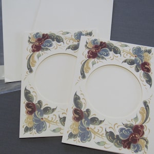 May include: Two cream-colored photo mats with floral designs in burgundy, blue, and gold. The mats have oval openings and are accompanied by blank envelopes, suitable for framing photos or creating personalized cards.