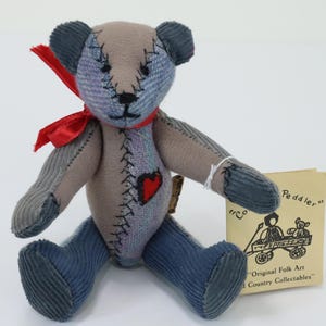May include: A stuffed teddy bear made of blue and grey corduroy fabric with visible black stitching, a red ribbon tied around its neck, and a red heart patch on its chest. A small beige tag is attached, featuring a drawing of a peddler and the text "Peddler" and "Original Folk Art Country Collectables".