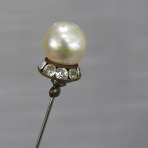 May include: A decorative stick pin featuring a round, off-white faux pearl topper. The base of the pearl is set in a dark metal cup adorned with a ring of small, clear, faceted crystals. The pin has a long, thin, silver-toned metal shaft.