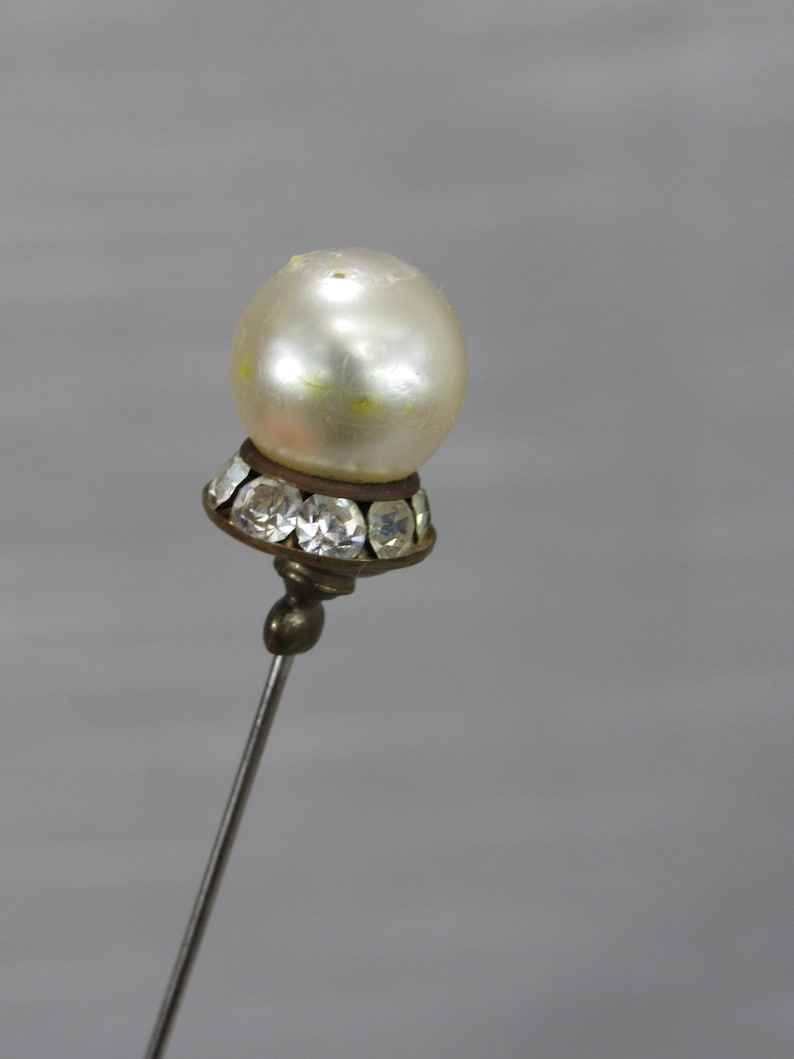 May include: A decorative stick pin featuring a round, off-white faux pearl topper. The base of the pearl is set in a dark metal cup adorned with a ring of small, clear, faceted crystals. The pin has a long, thin, silver-toned metal shaft.