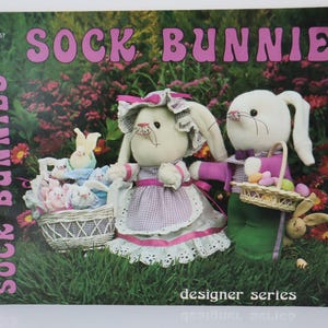 May include: A book cover with the title "SOCK BUNNIES" in pink letters. The cover features two plush bunnies, one wearing a dress and the other holding a basket of eggs. A basket of smaller bunnies is also visible.