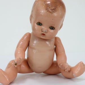 May include: A vintage composition doll with a molded peach-toned body, painted facial features including blue eyes and red lips, and sculpted hair. The doll is in a seated position with visible signs of wear, including scuffs and paint loss.