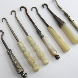 May include: A collection of six antique button hooks with varied handles. One features an engraved silver-tone metal handle, while the others have carved bone, smooth cream-colored, or iridescent mother-of-pearl handles with metal hooks.