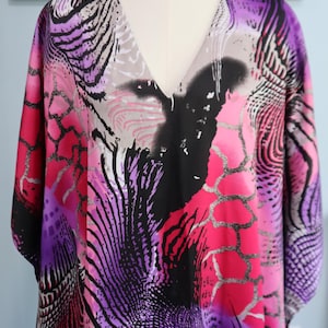 May include: A V-neck kaftan featuring a vibrant abstract print in shades of hot pink, purple, black, and white. The garment displays a bold, graphic pattern with swirling lines, organic shapes, and textured details across the entire fabric surface.