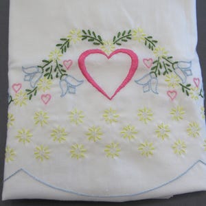 May include: A white fabric textile featuring embroidered designs including a central pink heart, light blue bell-shaped flowers, green leafy vines, and small yellow star-shaped blossoms. The bottom edge has a scalloped border with blue stitching.
