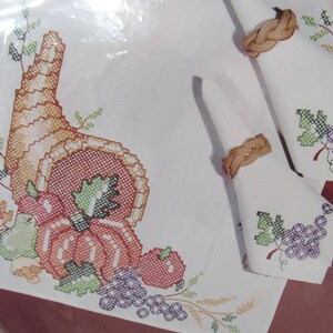 May include: A cross-stitch harvest-themed set featuring a cornucopia, pumpkins, grapes, and wheat. The set includes a placemat and two napkins with woven napkin rings. The placemat and napkins are white with colorful embroidery.