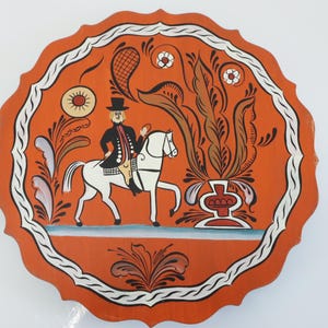 May include: Round, orange decorative plate with a white border and scalloped edge. The plate features a painted scene of a person on a white horse, with floral and foliate designs. The person is wearing a black coat and hat.