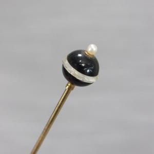 May include: A decorative hat pin featuring a long, slender gold-toned metal shaft topped with a spherical black bead. The bead is accented by a horizontal clear, translucent band around its center and is finished with a small white pearl at the top.