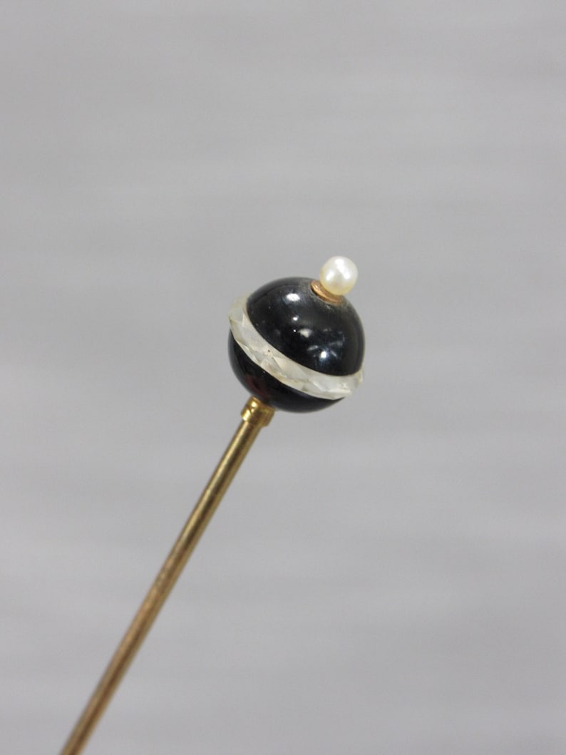 May include: A decorative hat pin featuring a long, slender gold-toned metal shaft topped with a spherical black bead. The bead is accented by a horizontal clear, translucent band around its center and is finished with a small white pearl at the top.