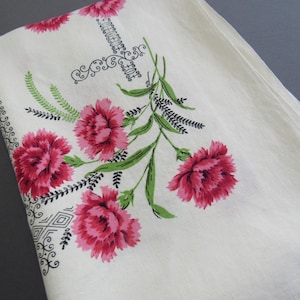 May include: A white fabric tablecloth featuring a printed floral pattern of vibrant pink carnations with green stems and leaves. The design includes black decorative scrollwork and geometric line borders along the edges of the textile.