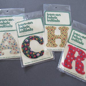 May include: Four individual packages of iron-on fabric initials, each containing a single letter: A, C, H, and R. Each letter features a distinct floral pattern in different colors, including light blue, dark blue, yellow, and red backgrounds.