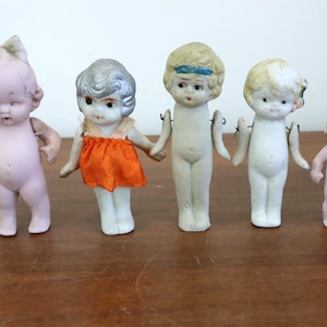 May include: Five antique bisque dolls with painted features and articulated arms. The dolls vary in size and wear different hairstyles and clothing. The dolls are arranged on a wooden surface.