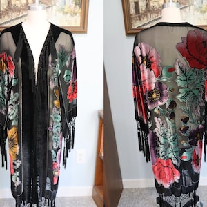 May include: Black kimono-style robe with a floral design in red, pink, yellow, and purple. The robe has a sheer overlay and velvet floral accents. It features long sleeves and fringe trim along the hem and sleeves. The robe is displayed on a mannequin.