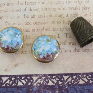 May include: Two round, hand-painted porcelain earrings with a floral design in shades of blue and white, set in gold-toned metal. A vintage brass thimble is also present, showcasing intricate detailing.