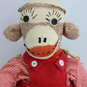 May include: A sock monkey doll featuring a cream and brown knit body, black button eyes, and a red yarn pom-pom on its head. The doll wears red corduroy overalls over a red and white striped long-sleeve shirt with white buttons.