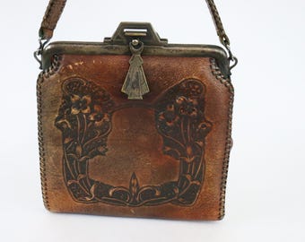May include: A vintage brown leather handbag with a decorative floral design. The bag features a metal clasp and a braided strap. The leather has a distressed appearance, adding to its antique charm. The bag is square-shaped.
