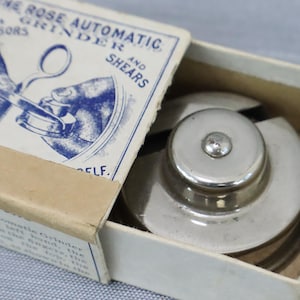 May include: A silver-toned metal Rose Automatic Grinder for scissors and shears housed in a white cardboard box. The box lid features blue printed text and a graphic illustration of the tool in use, with additional instructional text on the side.
