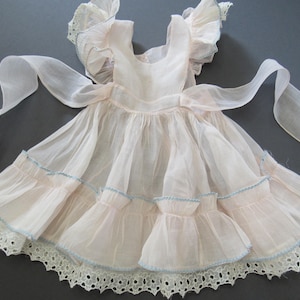 May include: A pale pink sleeveless dress featuring sheer ruffled shoulder straps, a gathered waist with long side ties, and a tiered skirt. The skirt layers are accented with light blue trim and finished with a white eyelet lace hem border.