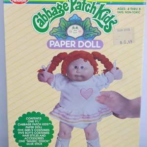 May include: Cabbage Patch Kids paper doll box. The box features a doll with red pigtails, wearing a white dress with a pink heart. The box includes the text "PAPER DOLL" and "AGES: 4 THRU 8". The box is 9.5 inches tall.