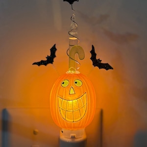 May include: An orange pumpkin-shaped night light with a glowing interior, featuring a wide smiling face with black outlines. The light is topped with a clear spiral stem, a yellow curved accent, and three black glittery bat cutouts attached to wires.