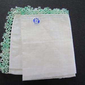 May include: White linen handkerchief with a delicate green lace trim. The handkerchief has a blue and white "Irish Linen" pin attached. The lace trim is a light green color and is along one edge of the handkerchief.