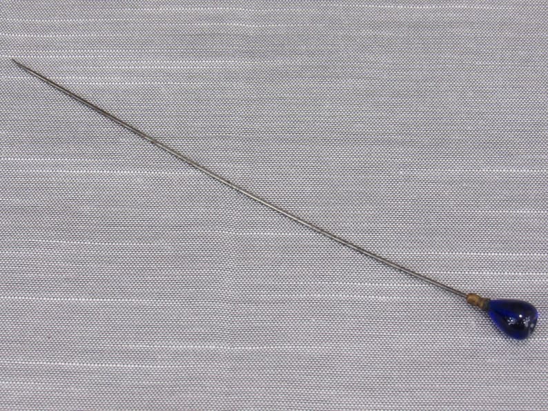 May include: A long, slender metal hat pin featuring a pointed tip and a smooth, teardrop-shaped dark blue glass bead at the opposite end. The pin has a metallic silver-toned finish and a small gold-toned metal cap connecting the bead to the shaft.